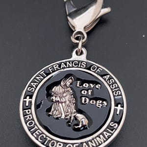Puede incluir: Un dije plateado con un cierre de mosquet&oacute;n. El dije presenta un centro negro con una imagen de San Francisco de As&iacute;s y un perro, con las palabras "Love of Dogs" y "Protector of Animals".