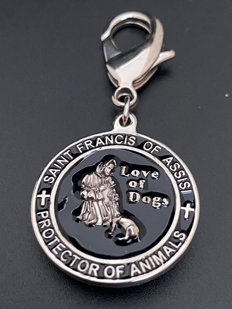 Puede incluir: Un dije plateado con un cierre de mosquet&oacute;n. El dije presenta un centro negro con una imagen de San Francisco de As&iacute;s y un perro, con las palabras "Love of Dogs" y "Protector of Animals".
