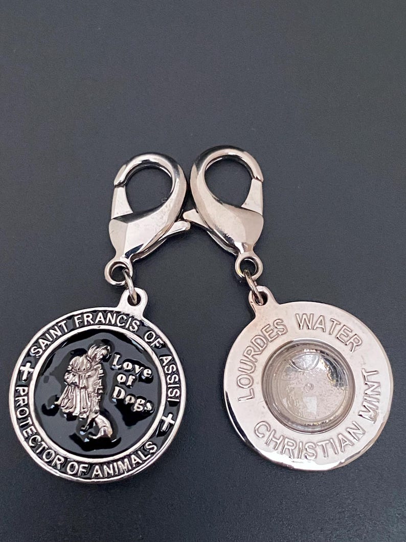 Puede incluir: Dos colgantes plateados con forma de chapa de perro. Uno presenta a San Francisco de As&iacute;s con el texto "Love of Dogs" y "Protector of Animals". El otro colgante tiene "Lourdes Water Christian Mint" alrededor de un centro transparente.