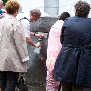 Puede incluir: Un grupo de personas alrededor de una fuente de agua. Una persona con un abrigo beige claro est&aacute; cerca de la fuente. Alguien est&aacute; llenando una botella de pl&aacute;stico transparente con agua.