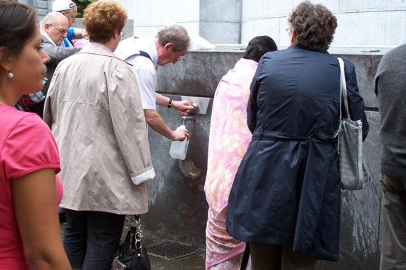 Puede incluir: Un grupo de personas alrededor de una fuente de agua. Una persona con un abrigo beige claro est&aacute; cerca de la fuente. Alguien est&aacute; llenando una botella de pl&aacute;stico transparente con agua.