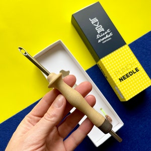 Op de afbeelding: Een houten handvatte punchenaald met een metalen naaldpunt. De naald zit in een witte doos met een gele en zwarte doos erachter. De doos heeft de tekst "Tricot Crochet" en "Punch Needle" erop.