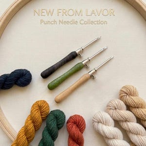 Op de afbeelding: Drie punch-naalden met houten handvatten in zwart, groen en naturel hout, diagonaal gerangschikt op een lichte stof in een houten borduurring. De tekst NEW FROM LAVOR Punch Needle Collection staat erboven. Verschillende garenstrengen in marineblauw, mosterdgeel, donkergroen, roestbruin, crème en beige omringen de naalden.