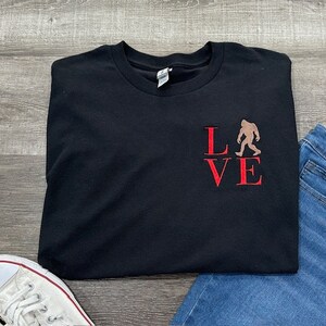May include: Black t-shirt with a red "LOVE" graphic featuring a silhouette of Bigfoot in the "O".