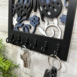 May include: Black wooden key holder with a butterfly and floral design. The key holder has six hooks and is hanging on a white wall.