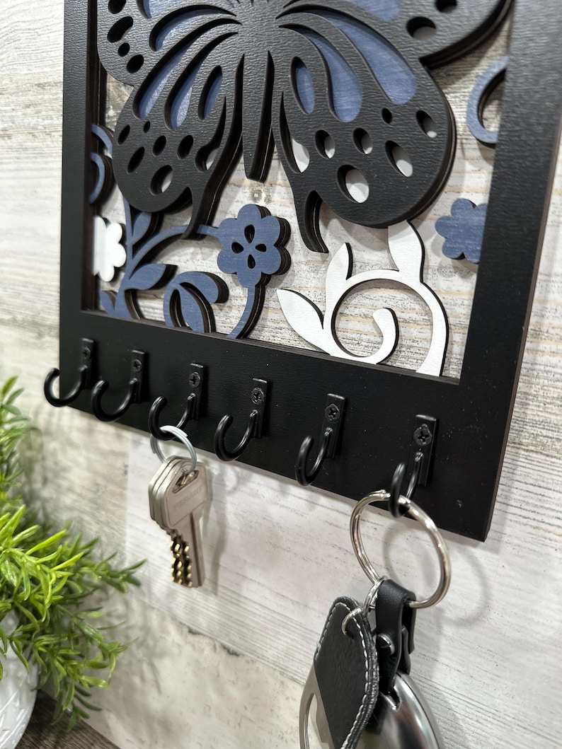 May include: Black wooden key holder with a butterfly and floral design. The key holder has six hooks and is hanging on a white wall.