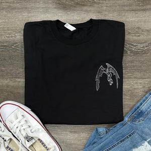 May include: Black t-shirt with a white embroidered design of a flying creature with wings. The creature has a small eye and a long tail.