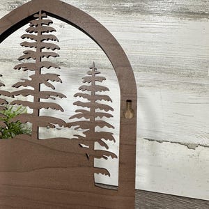 May include: A wooden wall hanging with a cutout design of three pine trees in a forest scene. The hanging has a brown stain and a hole for hanging on the right side.