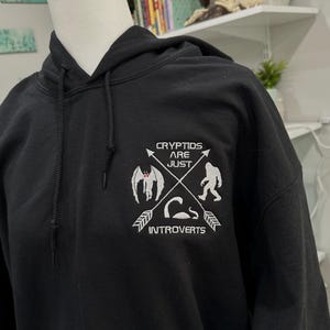 May include: Black hooded sweatshirt with a white embroidered design featuring a variety of mythical creatures and the text "Cryptids are just introverts".