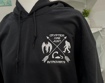 May include: Black hooded sweatshirt with a white embroidered design featuring a variety of mythical creatures and the text "Cryptids are just introverts".