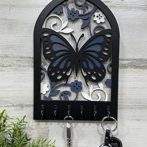 May include: Black wooden key holder with a blue and white butterfly design and six hooks. The key holder is shaped like an arch and has a floral pattern.