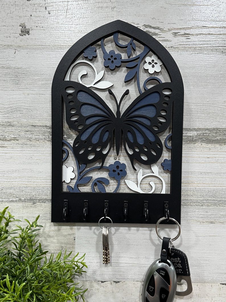 May include: Black wooden key holder with a blue and white butterfly design and six hooks. The key holder is shaped like an arch and has a floral pattern.