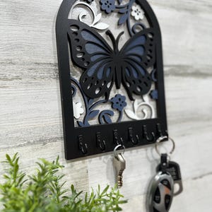 May include: Black wooden key holder with a blue and white butterfly design. The holder has six hooks for hanging keys.