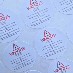 May include: Circular white warning labels with red text and a red exclamation mark symbol. The labels warn about flammable liquid, not lighting reeds, and keeping out of reach of children and pets. The text also provides usage instructions.