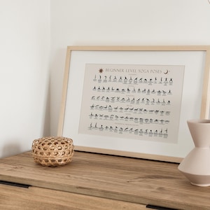 May include: A framed print of a beginner level yoga poses chart with black and white illustrations. The chart is on a light beige background with a sun and moon symbol at the top. The print is on a wooden dresser with two vases and a woven basket.