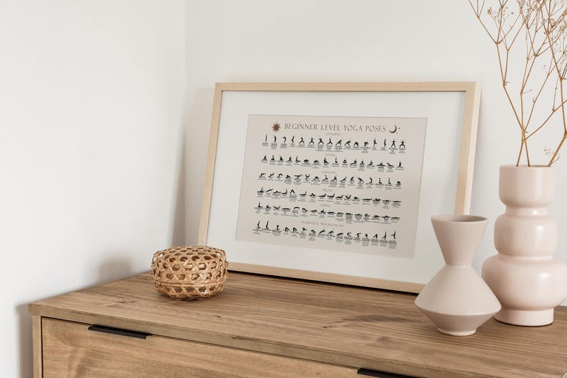 May include: A framed print of a beginner level yoga poses chart with black and white illustrations. The chart is on a light beige background with a sun and moon symbol at the top. The print is on a wooden dresser with two vases and a woven basket.