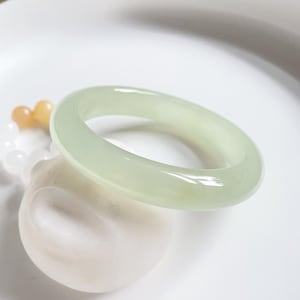 May include: A light green jade bangle bracelet. The bangle is smooth and polished, and it has a slightly rounded shape.