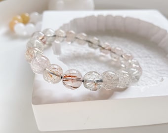 May include: A clear quartz bracelet with red and silver inclusions. The bracelet is made of round beads and is strung on a black elastic cord.