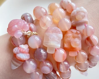 May include: A set of four bracelets made of pink and orange agate beads. The beads are round and have a natural, polished finish. Some of the beads are carved into animal shapes. The bracelets are stacked on a person's wrist.