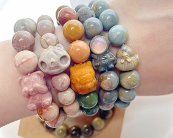 May include: A stack of colorful bracelets with various stone beads and small animal charms. The bracelets are made of different types of stones, including pink, blue, green, and brown. The charms are made of white and brown stone and feature animal designs, including a cat, a lion, a bird, and a bear.