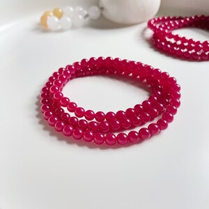 May include: A strand of pink gemstone beads, likely made of rose quartz or pink jade, is wrapped around itself in a circular shape. The beads are smooth and polished, and the color is a vibrant pink.
