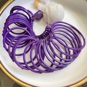 May include: A set of purple ring sizers arranged in a circular pattern. The sizers are made of plastic and are connected by a metal wire. The set is on a white plate, with a white decorative object and a beaded bracelet in the background.