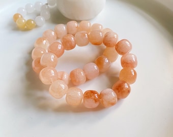 May include: A peach-colored gemstone bracelet with rounded beads. The bracelet is made of polished stones, with variations in color from light peach to a deeper orange. A white and yellow beaded necklace and a white decorative object are also visible.