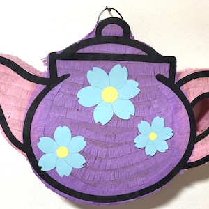 May include: A purple and pink teapot piñata with a black outline and three blue paper flowers with yellow centers.