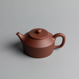 以下が含まれることがあります： 蓋と取っ手が付いた茶色のセラミックティーポット。 ティーポットは丸みを帯びたボディと注ぎ口を備えています。 蓋の上部には小さなノブが付いています。 ティーポットは灰色の背景に置かれています。