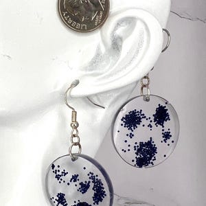May include: Clear, round earrings with silver hooks and dark blue glitter designs. The earrings are shown next to a U.S. dime for size comparison. The glitter is clustered in various shapes within the clear resin.
