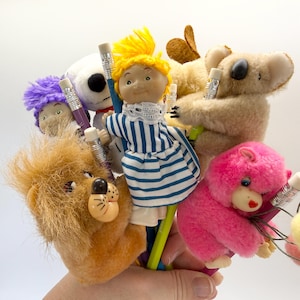 May include: A collection of pencil toppers featuring various plush characters including a doll with yellow yarn hair in a blue and white striped dress, a brown lion, a pink gorilla, a tan koala, a purple-haired figure, and a white character.