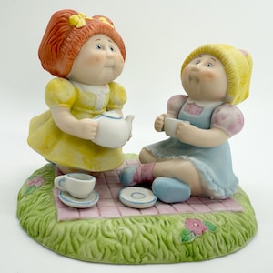 May include: A ceramic figurine depicting two dolls having a tea party. One doll wears a yellow dress and holds a teapot, while the other wears a blue dress and holds a teacup. The base is green with a pink checkered blanket and small tea set.