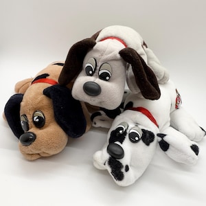 May include: Three plush puppy dog toys in various colors. One is tan with black ears, another is white with brown ears and spots, and the third is gray with brown ears. Each has a red collar and large, cartoonish eyes.