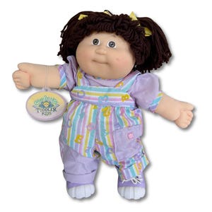 May include: A Cabbage Patch Kids Toddler doll with brown yarn hair in pigtails, wearing a lavender outfit with a striped pattern and the text "Cabbage Patch Kids Toddler Kids" on the tag.