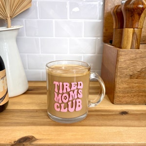 May include: A clear glass mug with a pink "TIRED MOMS CLUB" graphic filled with coffee.