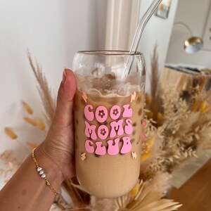 May include: A glass with a pink and white sticker that says "Cool Moms Club" with a flower design. The glass is filled with iced coffee and a clear glass straw is sticking out of the top.