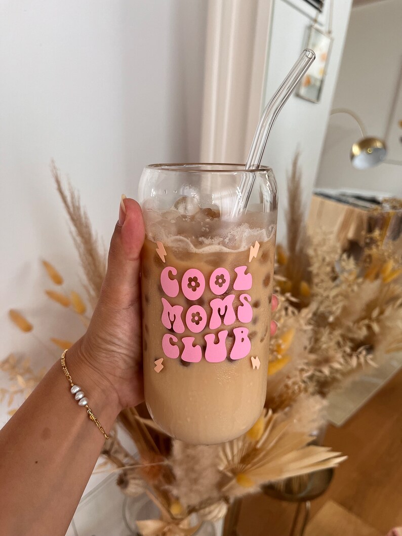 May include: A glass with a pink and white sticker that says "Cool Moms Club" with a flower design. The glass is filled with iced coffee and a clear glass straw is sticking out of the top.