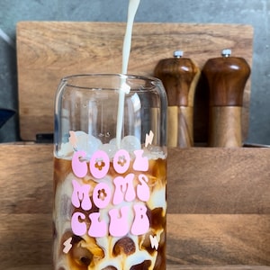 May include: A glass with ice and coffee is being poured with milk. The glass has a pink graphic that says "Cool Moms Club" with flowers.