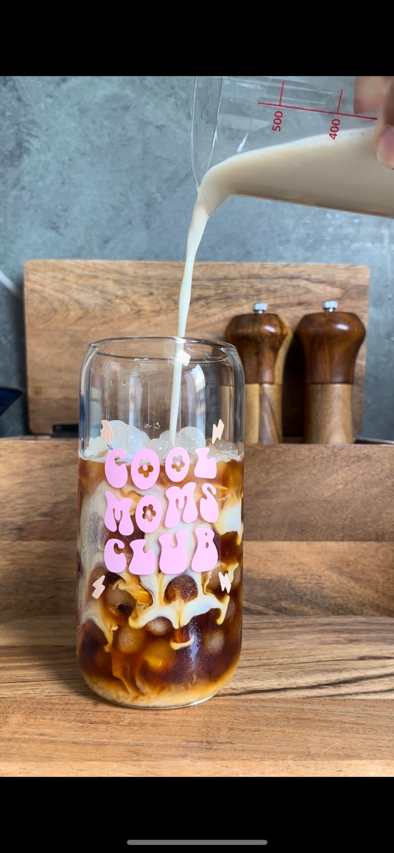 May include: A glass with ice and coffee is being poured with milk. The glass has a pink graphic that says "Cool Moms Club" with flowers.