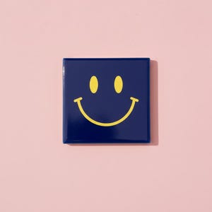 Könnte beinhalten: Eine marineblaue quadratische Fliese mit einem gelben Smiley-Gesicht-Design. Die Fliese hat eine glänzende Oberfläche und ist vor einem hellrosa Hintergrund platziert. Das Smiley-Gesicht hat zwei ovale Augen und einen gebogenen Mund.