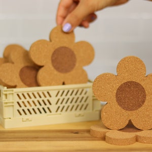 May include: A set of six cork flower-shaped coasters in a light beige crate. The coasters are brown with a darker brown center.