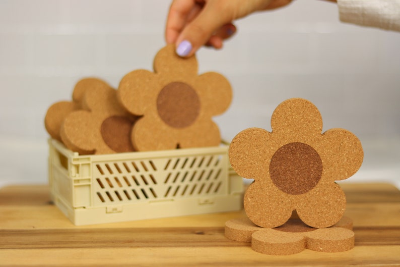 May include: A set of six cork flower-shaped coasters in a light beige crate. The coasters are brown with a darker brown center.