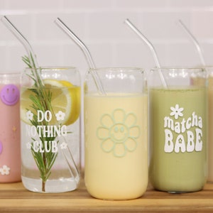 May include: Five glass cups with straws and different drinks. The cups have various designs, including a smiley face flower, the words "Do Nothing Club", and "Matcha Babe".