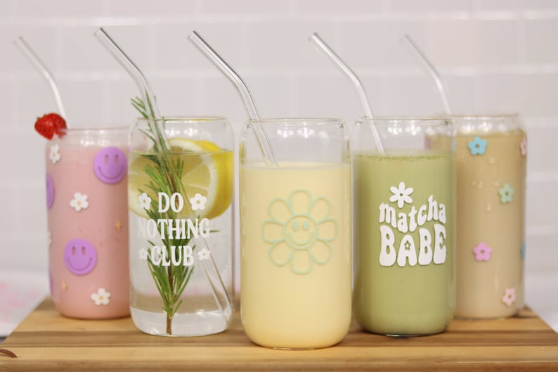 May include: Five glass cups with straws and different drinks. The cups have various designs, including a smiley face flower, the words "Do Nothing Club", and "Matcha Babe".