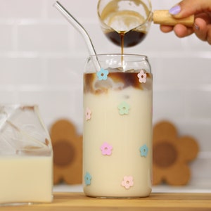 Könnte beinhalten: Ein Glas mit Eiskaffee, in das Milch aus einem kleinen Krug gegossen wird. Das Glas hat pastellfarbene rosa, blaue und gelbe Blumenaufkleber. Das Glas steht auf einer Holzoberfläche mit einem braunen Untersetzer.