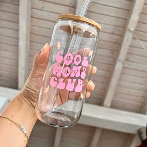 May include: A clear glass water bottle with a bamboo lid and a pink and white "Cool Moms Club" graphic. The bottle has a clear glass straw.