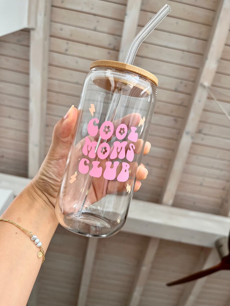 May include: A clear glass water bottle with a bamboo lid and a pink and white "Cool Moms Club" graphic. The bottle has a clear glass straw.