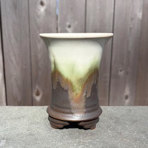 May include: A handcrafted ceramic vase with a unique color gradient. The top is a creamy white, transitioning to a soft green, then a rich brown at the base. The vase has a flared rim and sits on a small, decorative base.