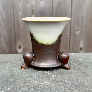 May include: A ceramic vase with a flared top and three small feet. The vase has a gradient of colors, starting with a light beige rim, transitioning to white, then green, and finally a dark brown base. The vase is on a gray surface.