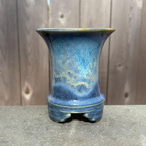 May include: A ceramic bonsai pot with a flared rim and a reactive glaze featuring shades of deep navy, royal blue, and light turquoise. The pot has a cylindrical body that tapers slightly toward the center and rests on four small feet.